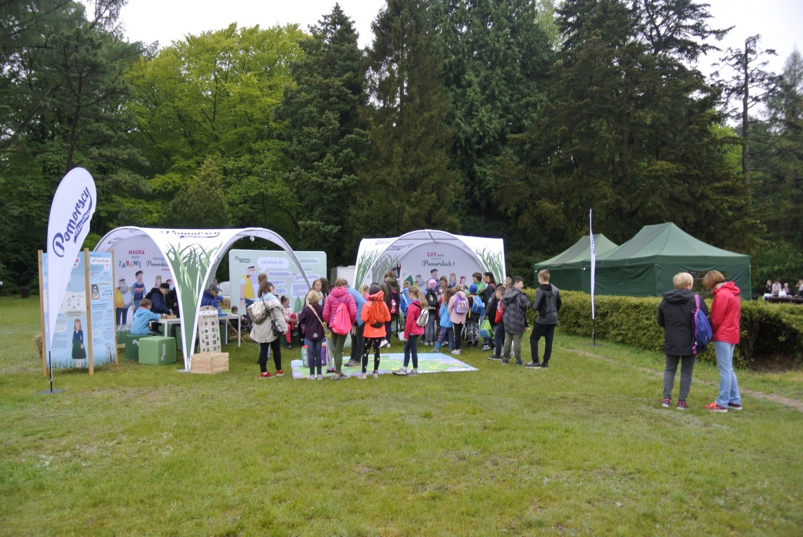 w centralnej części kadru widoczne jest stanowisko edukacyjne z dwoma namiotami kampanii „Poczuj klimat”, przy którym widać rozproszoną grupę dzieci, ponad 20 osób, wraz trzema opiekunami. Dwóch edukatorów prowadzi edukację.