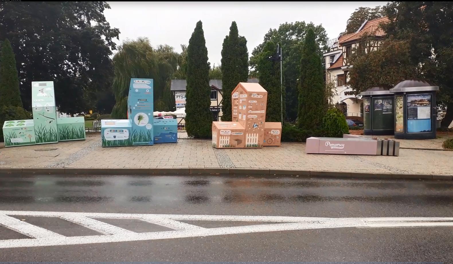 układ czterech, różnego koloru brył przestrzennych. Na trzech znajdują się treści edukacyjne, rysunki i diagramy. Bryły ustawione są w szeregu. Na najniższej bryle, będącej ławką, znajduje się logo kampanii „Pomorscy czują klimat”