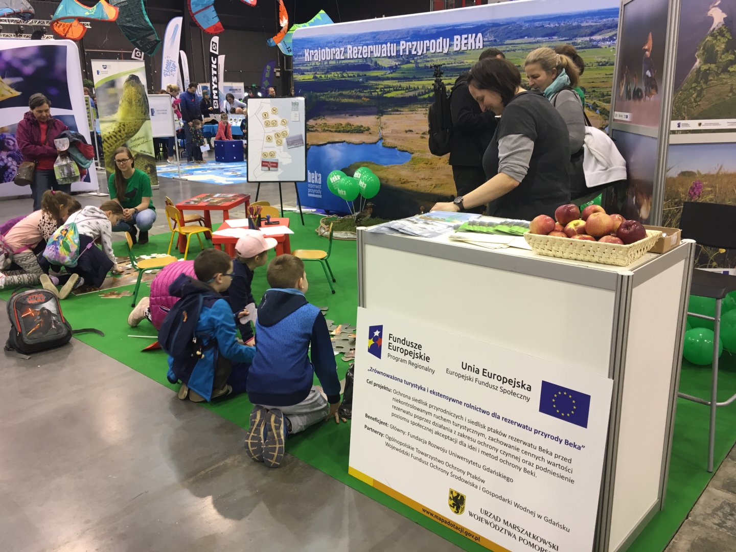 Przy stoisku ekspozycyjnym widać tablicę informacyjną z tytułem projektu „Zrównoważona turystyka i ekstensywne rolnictwo dla rezerwatu przyrody Beka”. W głębi znajdują się dzieci składające układanki z podobiznami ptaków 