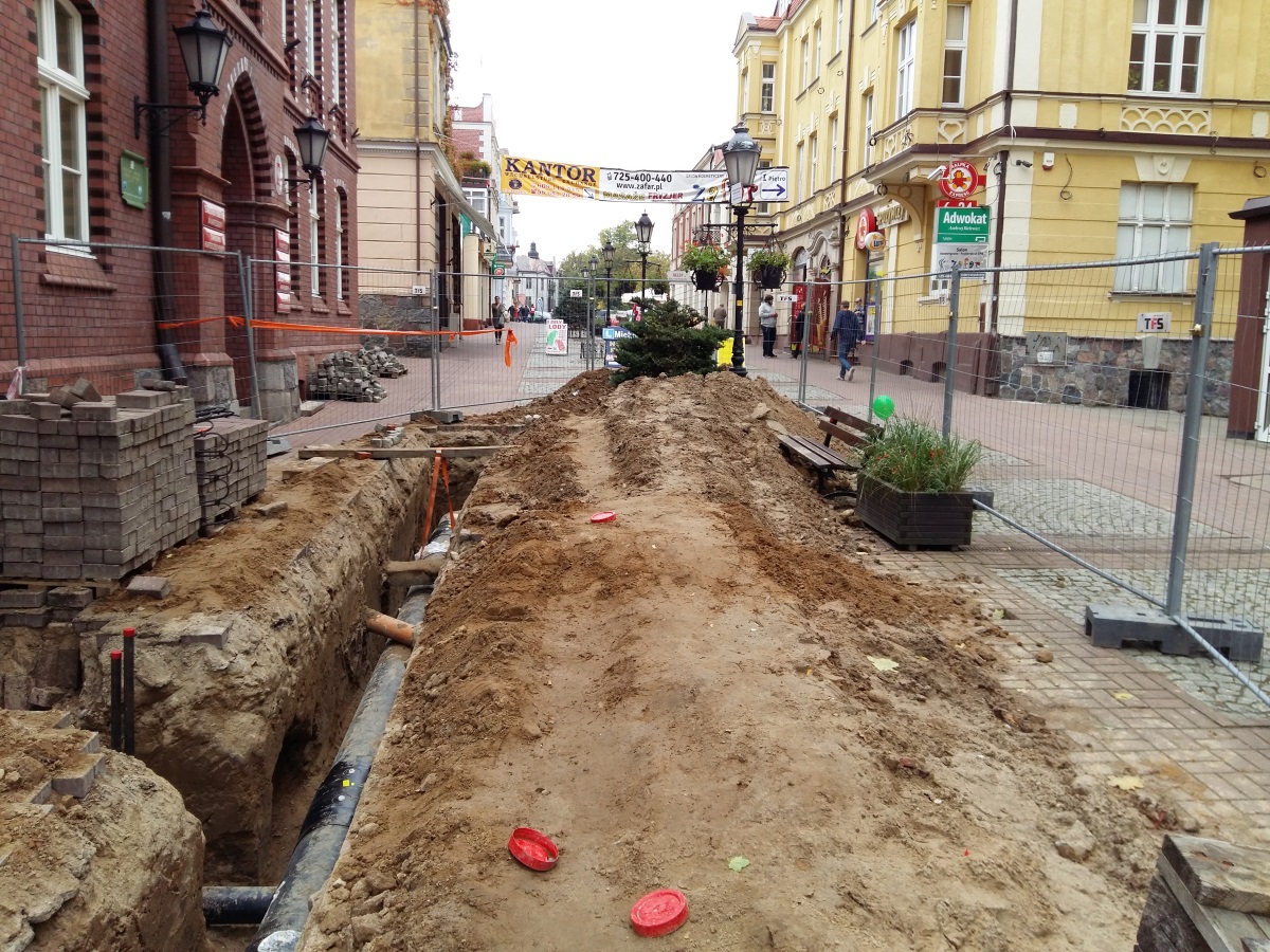 Budowa sieci ciepłowniczej w Kartuzach; centrum miasta, deptak, po obu stronach budynki, wzdłuż deptaku rów, w środku rura ciepłownicza 