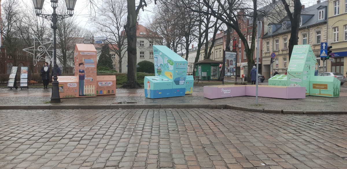 Interaktywna wystawa plenerowa, składająca się z trzech brył i ławki, na placu w Słupsku