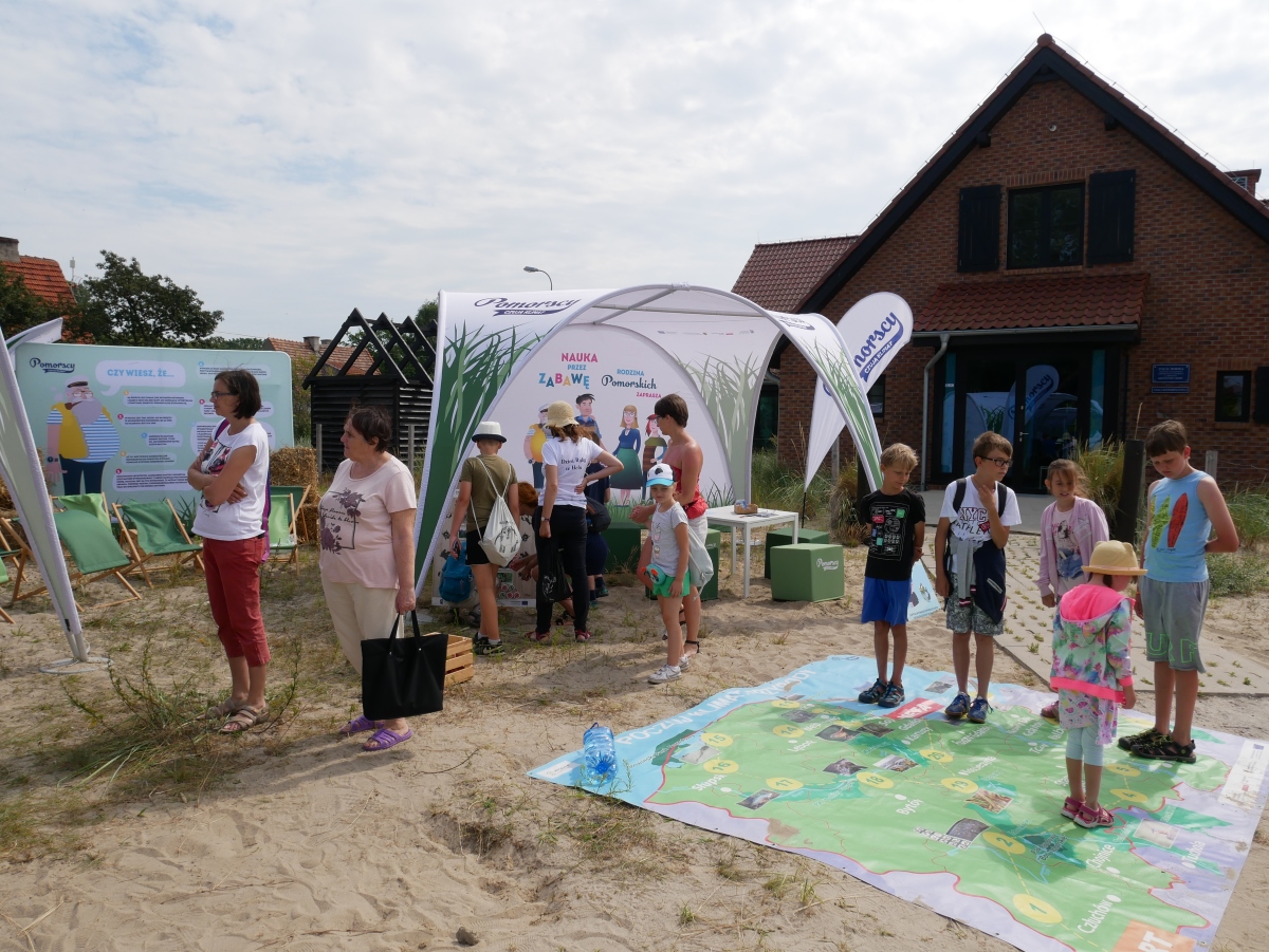 Edukacyjne stoisko Rodziny Pomorskich podczas Dnia Ryby w Helu; na zdjęciu widać namiot, na ziemi leżą rozłożone puzzle, dookoła dużo dzieci i dorosłych