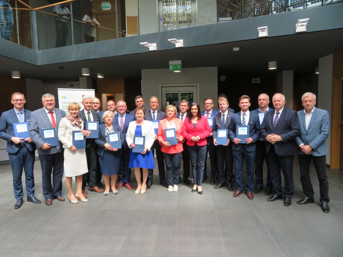 Na zdjęciu widać grupę osób, część z nich trzyma w rękach granatowe teczki z logo programu "Czyste Powietrze"