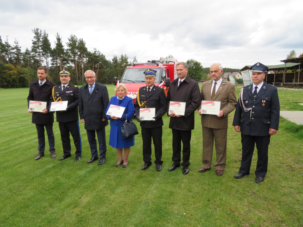 Na boisku w rzędzie stoją uczestnicy uroczystości przekazania samochodu ratowniczo-gaśniczego dla OSP Piece, za nimi widać czerwony samochód pożarniczy
