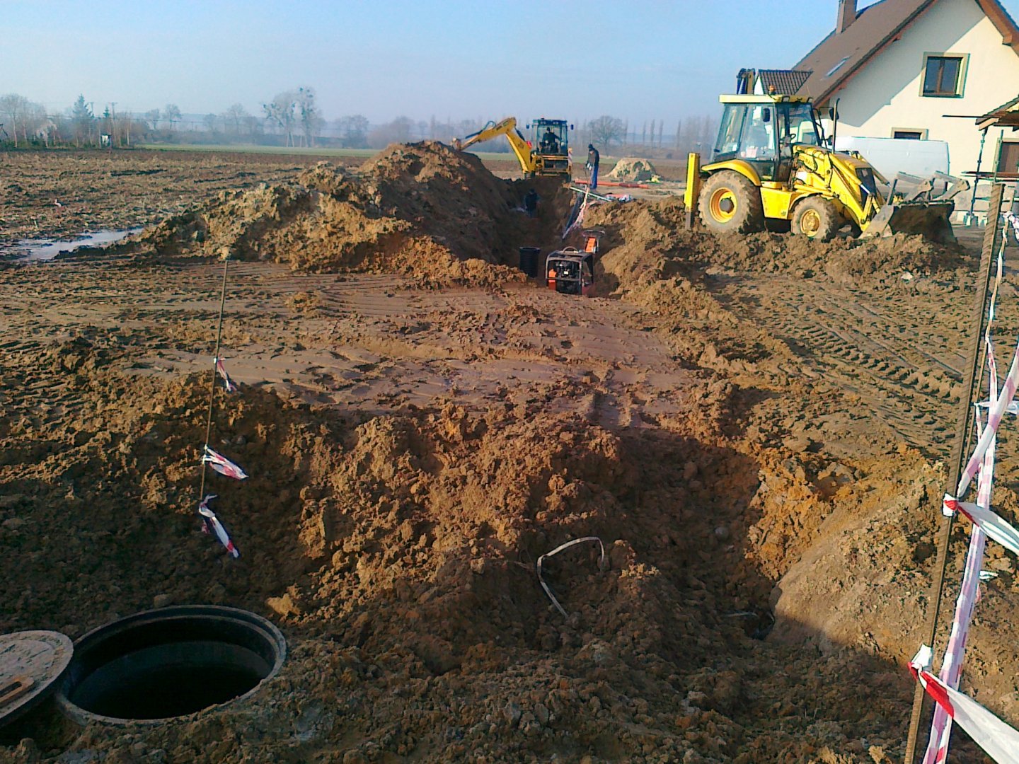 Budowa kanalizacji sanitarnej w miejscowości Rakowiec – rozkopana ziemia obok domu mieszkalnego, w ziemię wkopane zbiorniki, na dalszym planie koparka