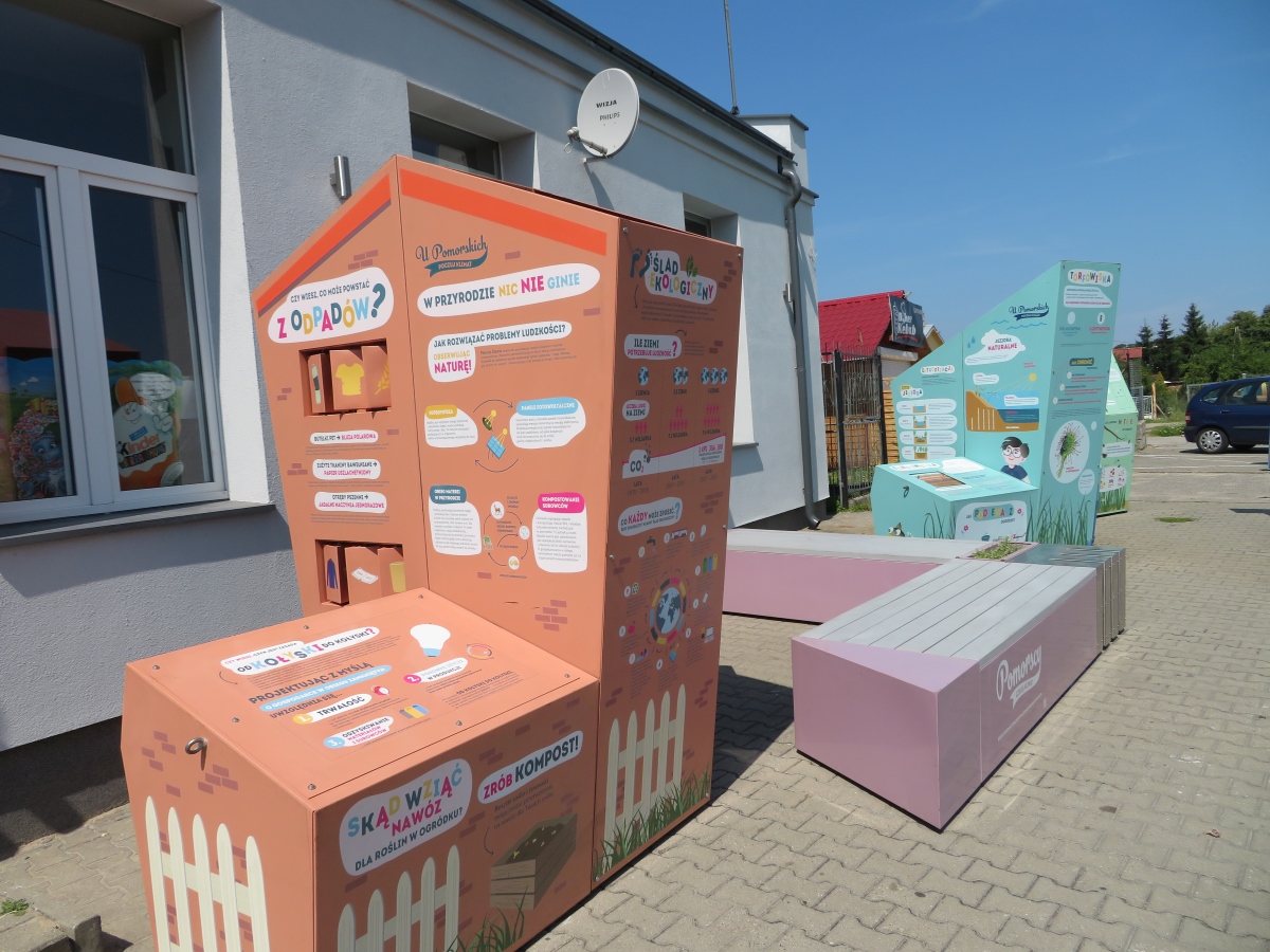 Na zdjęciu widać trzy kolorowe bryły oraz ławkę. Całość tworzy interaktywną wystawę plenerową