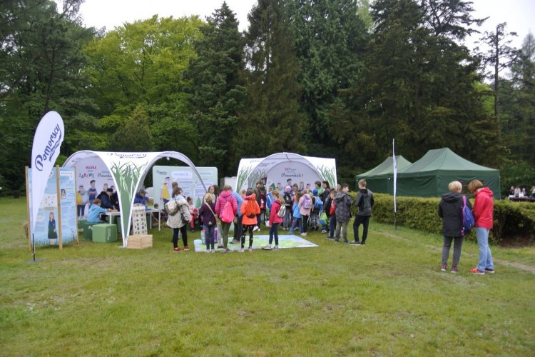 w centralnej części kadru widoczne jest stanowisko edukacyjne z dwoma namiotami kampanii „Poczuj klimat”, przy którym widać rozproszoną grupę dzieci, ponad 20 osób, wraz trzema opiekunami. Dwóch edukatorów prowadzi edukację.