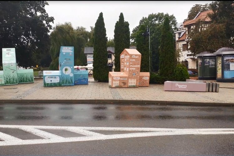 układ czterech, różnego koloru brył przestrzennych. Na trzech znajdują się treści edukacyjne, rysunki i diagramy. Bryły ustawione są w szeregu. Na najniższej bryle, będącej ławką, znajduje się logo kampanii „Pomorscy czują klimat”