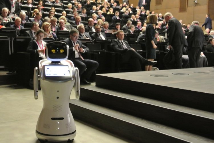 na pierwszym planie widoczny jest stojący robot humanoidalny. W tle siedzące osoby z publiczności.