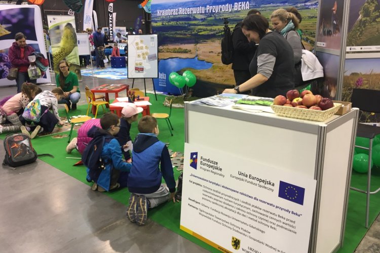 Przy stoisku ekspozycyjnym widać tablicę informacyjną z tytułem projektu „Zrównoważona turystyka i ekstensywne rolnictwo dla rezerwatu przyrody Beka”. W głębi znajdują się dzieci składające układanki z podobiznami ptaków 