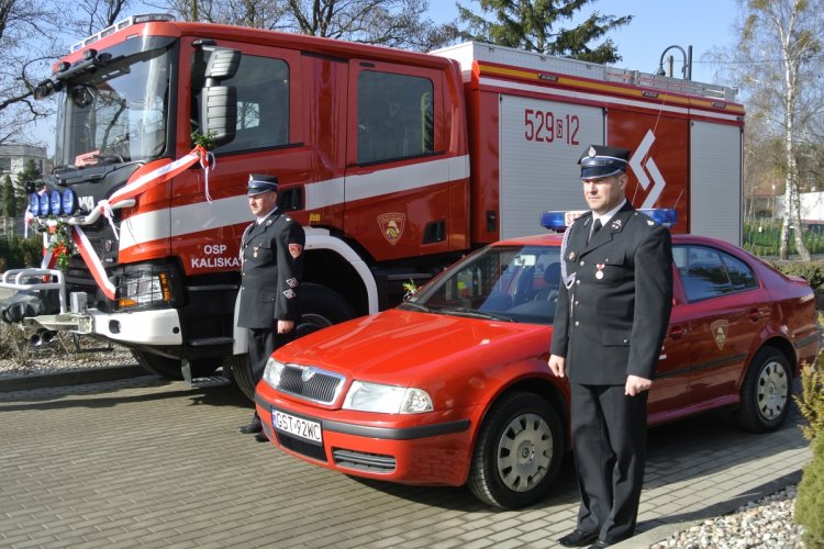 Ciężki, czerwony samochód ratowniczo-gaśniczy, obok stoi mały, czerwony samochód, po bokach strażacy w mundurach