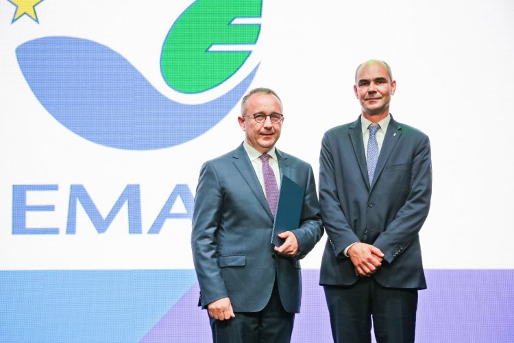 Na zdjęciu widać dwóch mężczyzn, jeden z nich trzyma w ręku teczkę, z tyłu napis EMAS.