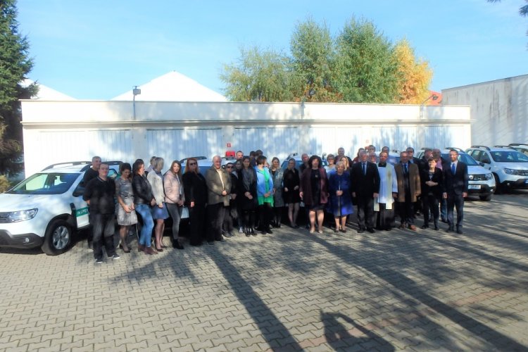 Na placu stoi grupa osób za nimi białe  samochody.