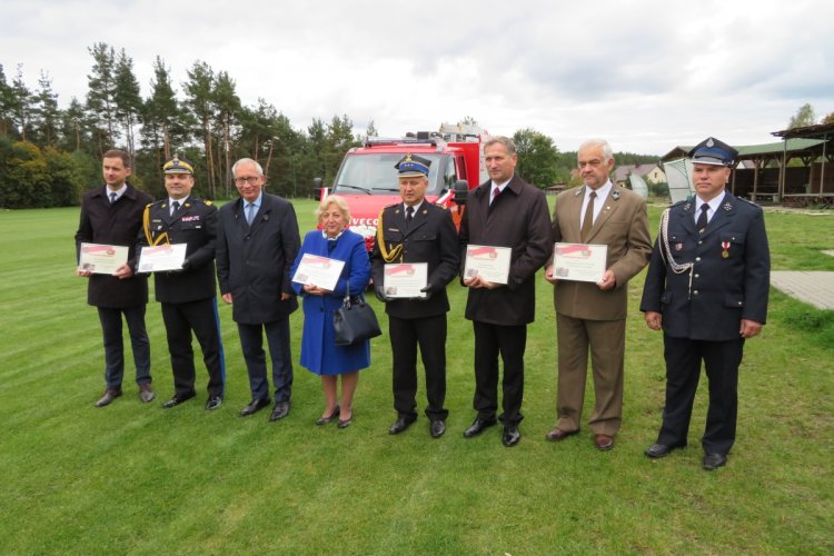 Na boisku w rzędzie stoją uczestnicy uroczystości przekazania samochodu ratowniczo-gaśniczego dla OSP Piece, za nimi widać czerwony samochód pożarniczy