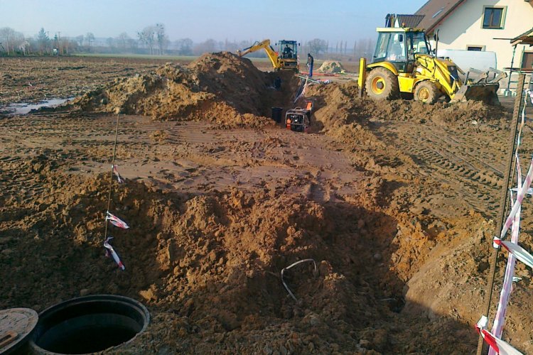 Budowa kanalizacji sanitarnej w miejscowości Rakowiec – rozkopana ziemia obok domu mieszkalnego, w ziemię wkopane zbiorniki, na dalszym planie koparka