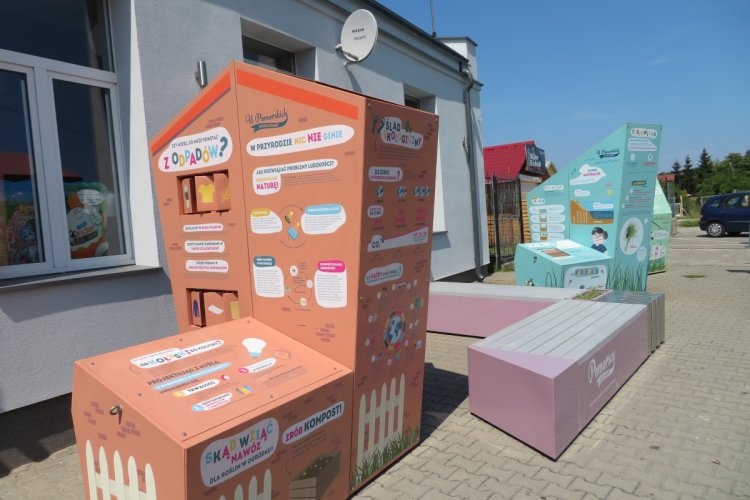 Na zdjęciu widać trzy kolorowe bryły oraz ławkę. Całość tworzy interaktywną wystawę plenerową